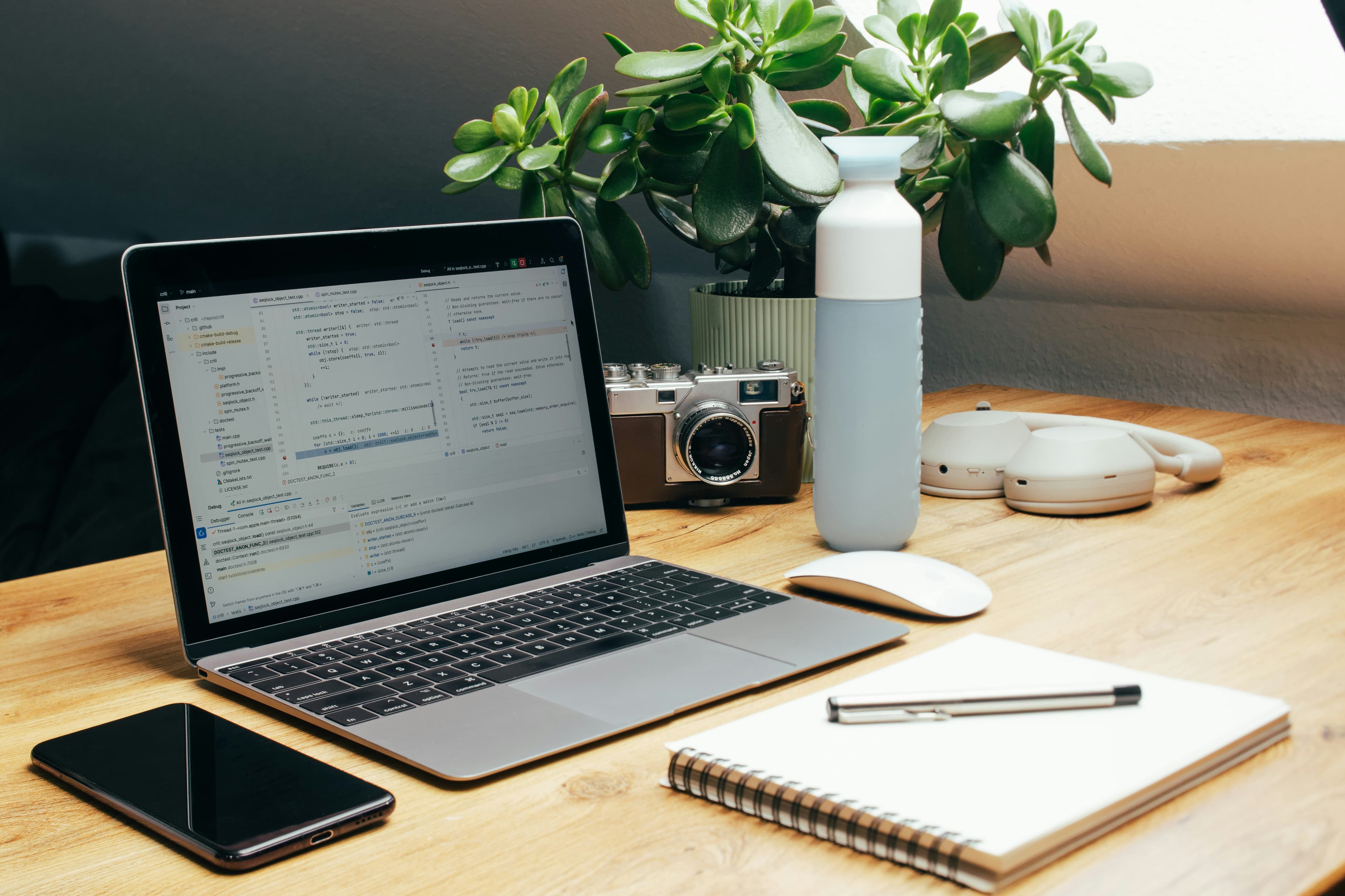 A neatly organized wooden desk features a laptop, a smartphone, a notebook, and various tech accessories next to a green potted plant.