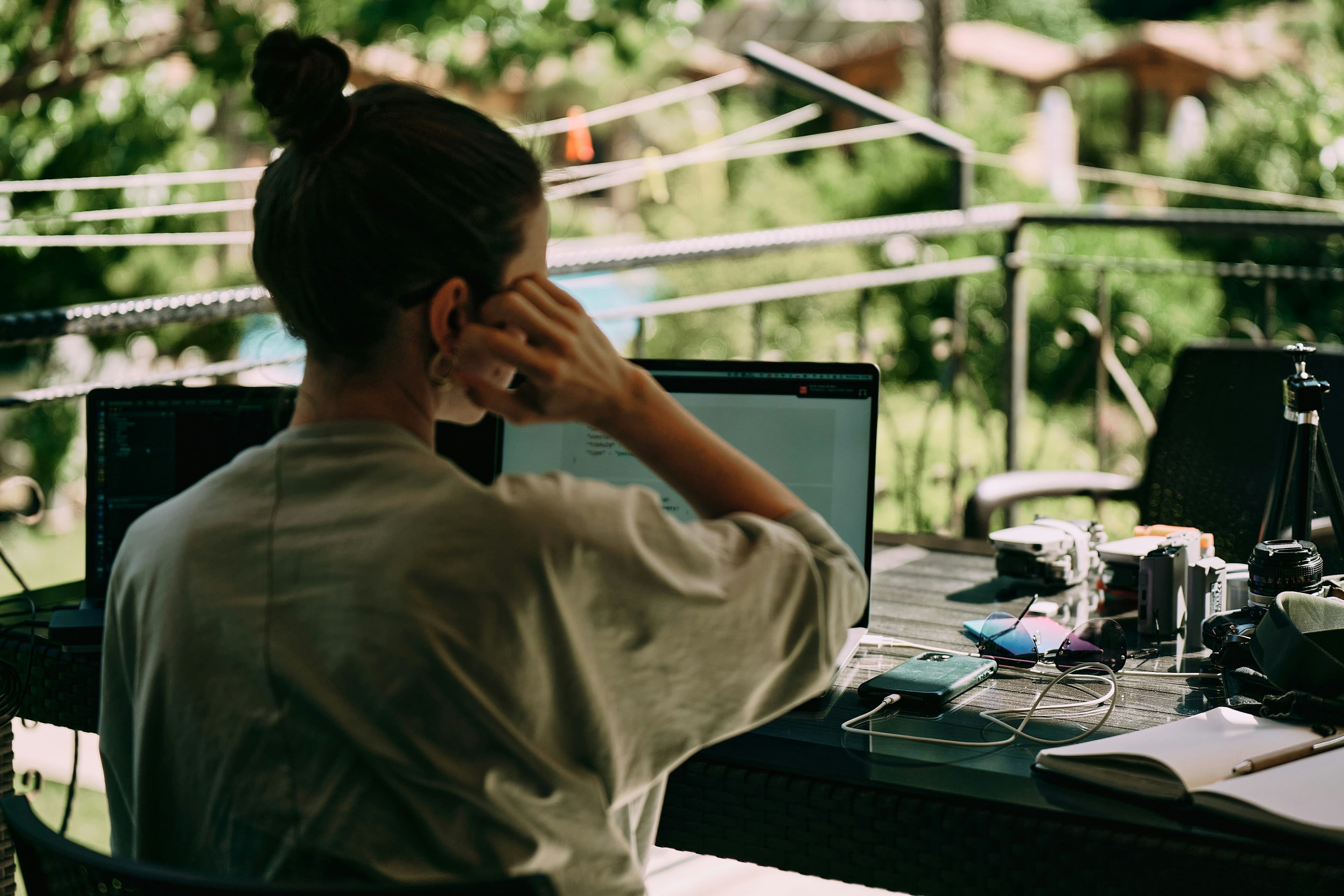 A woman works on her laptop at an outdoor table surrounded by lush greenery.