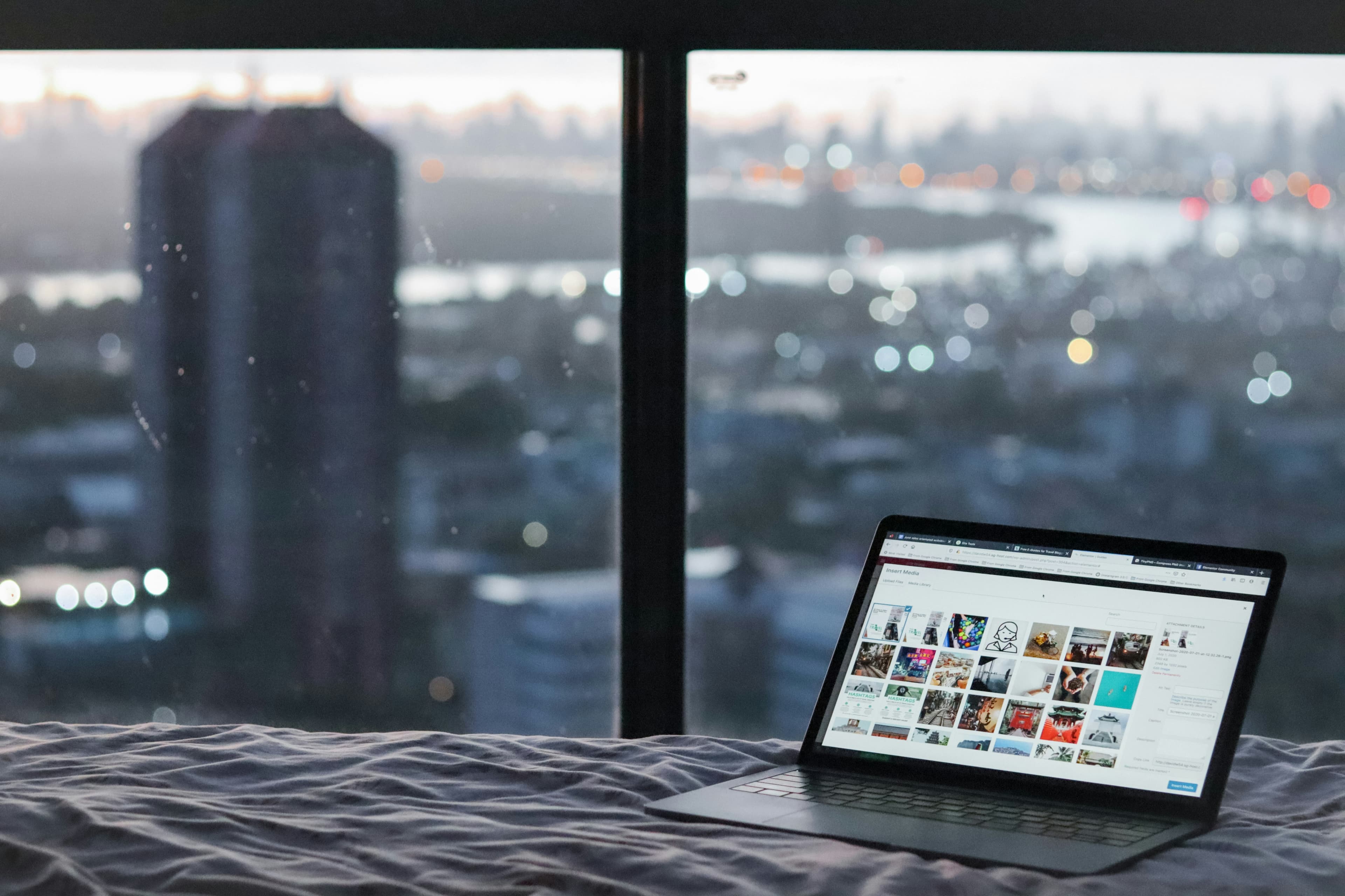 A laptop sits on a bed in front of a window overlooking a blurred city skyline at dusk.